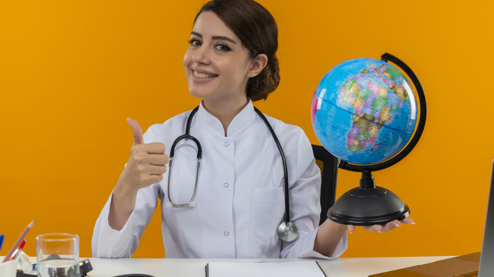 smiling young female doctor wearing medical robe and stethoscope sitting at desk with medical tools and laptop holding globe showing thumb up isolated on yellow background