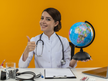 smiling young female doctor wearing medical robe and stethoscope sitting at desk with medical tools and laptop holding globe showing thumb up isolated on yellow background