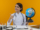 smiling young female doctor wearing medical robe and stethoscope sitting at desk with medical tools and laptop holding globe showing thumb up isolated on yellow background