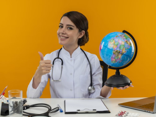 smiling young female doctor wearing medical robe and stethoscope sitting at desk with medical tools and laptop holding globe showing thumb up isolated on yellow background